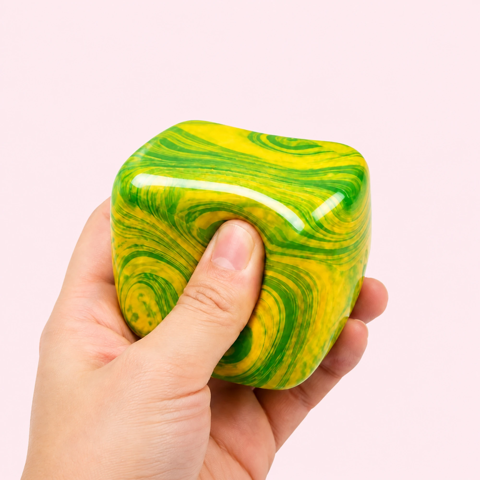 Hand squeezing the green and yellow Fluid Swirl Cube on a pale pink background