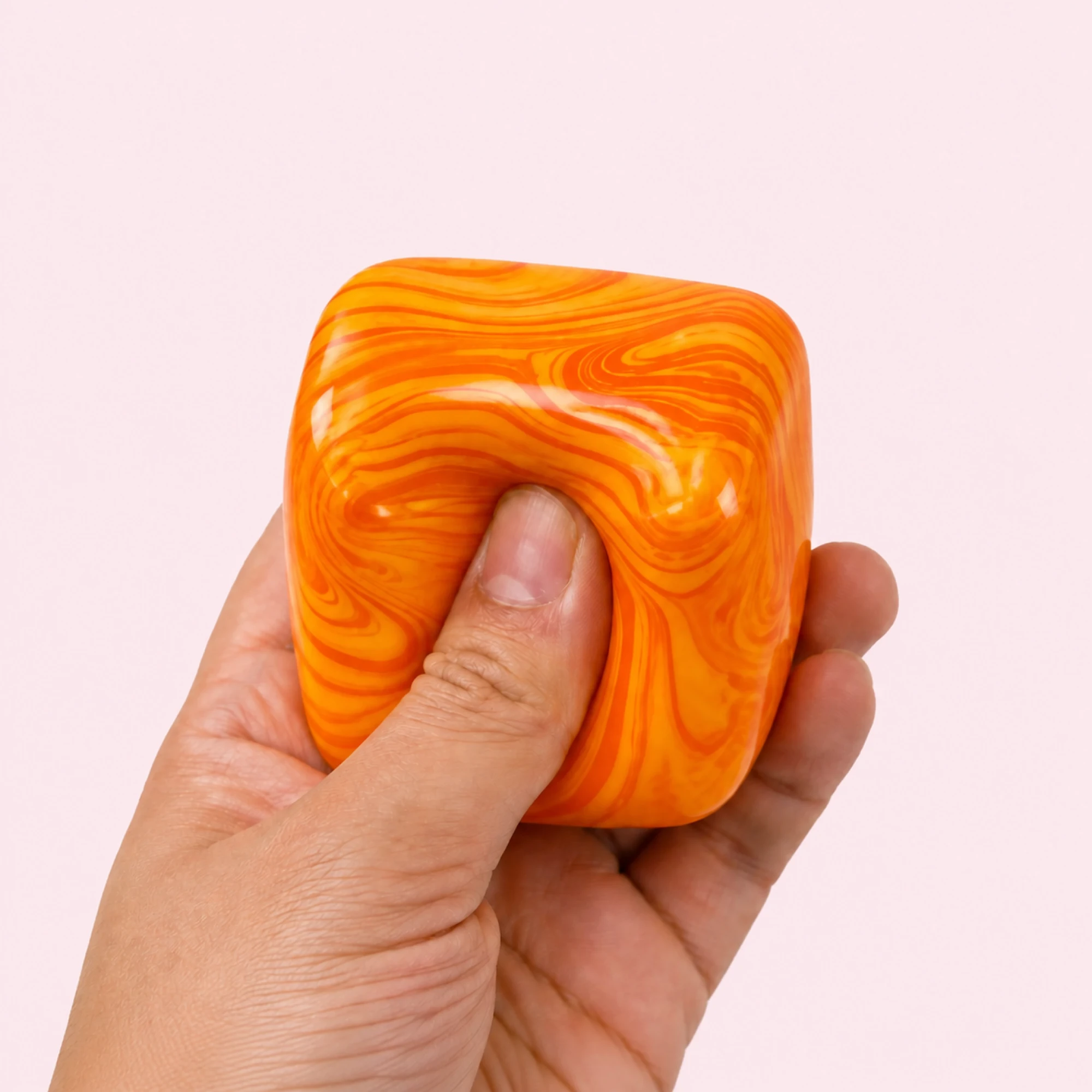 Hand squeezing the orange and yellow Fluid Swirl Cube on a pale pink background